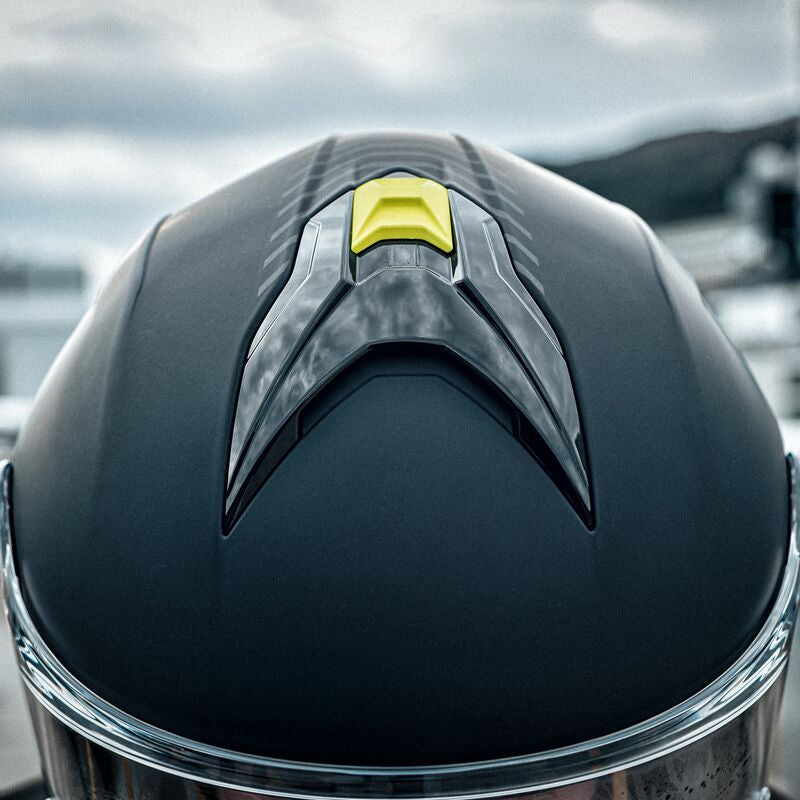 Close-up of a black motorcycle helmet with a glossy finish and a small bright yellow accent on top, photographed outdoors with a blurred racetrack or industrial background.
