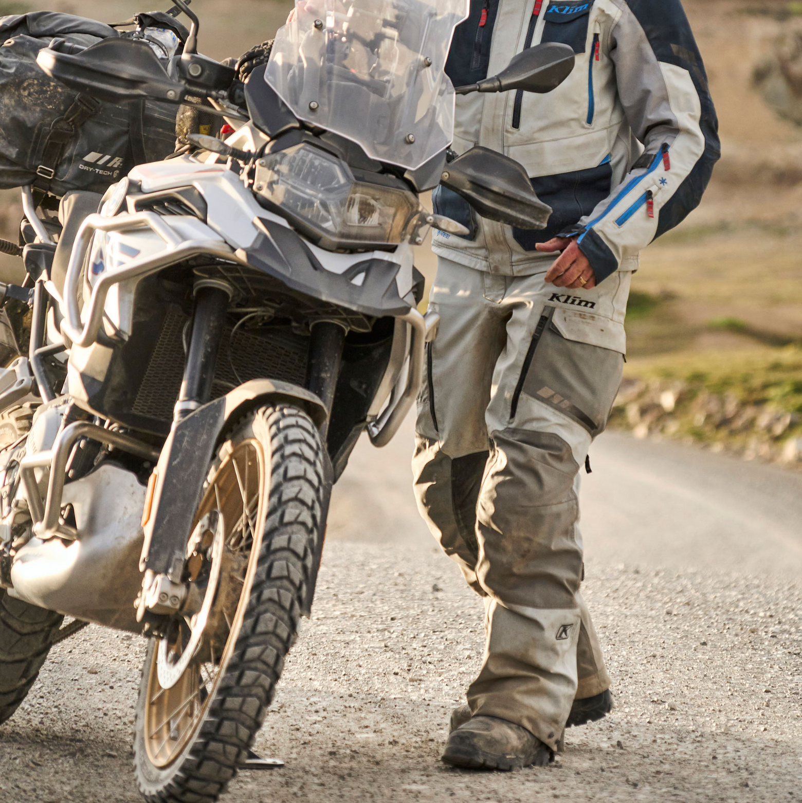 Motorcyclist in protective gear walking beside a motorcycle on a dirt road with mountains in the background