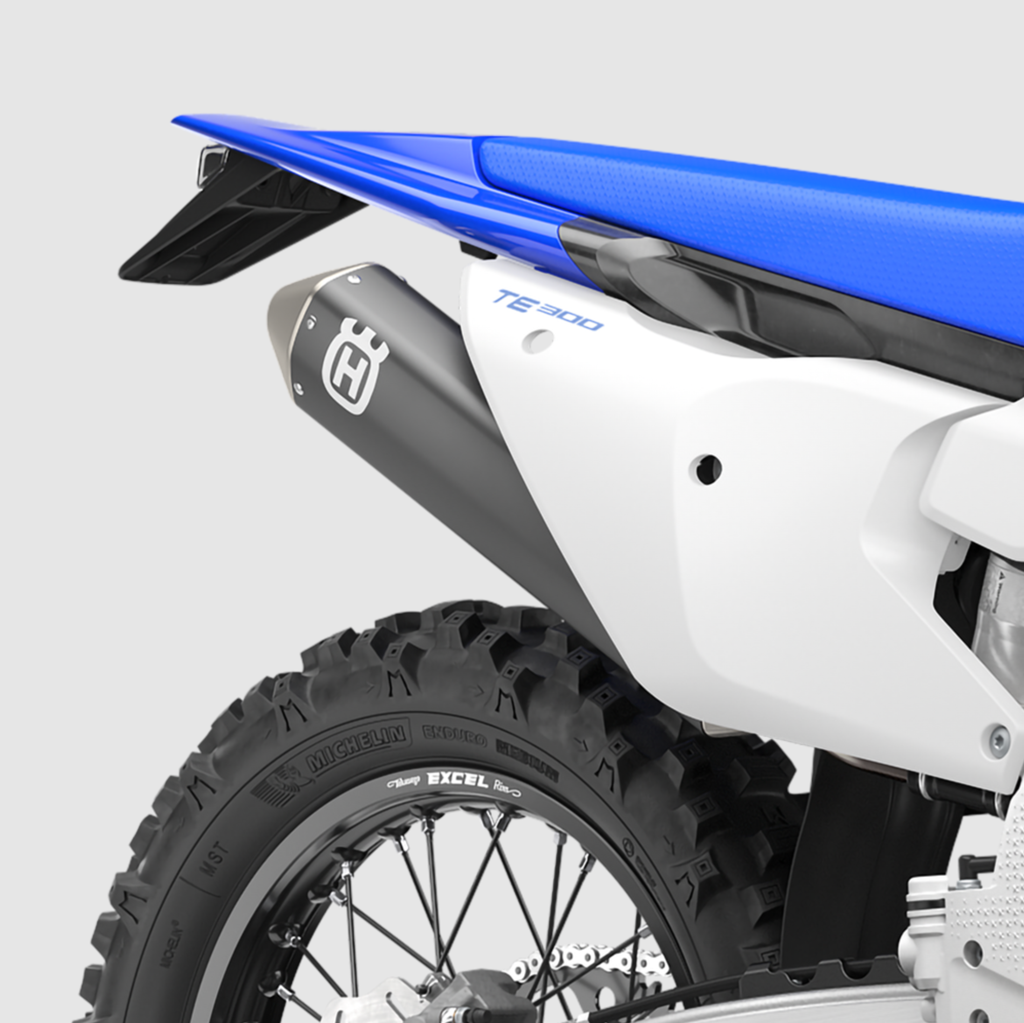 Close-up of a white and blue motorcycle rear section with a black Husqvarna exhaust muffler, rear tire, and blue seat on a light gray studio background.