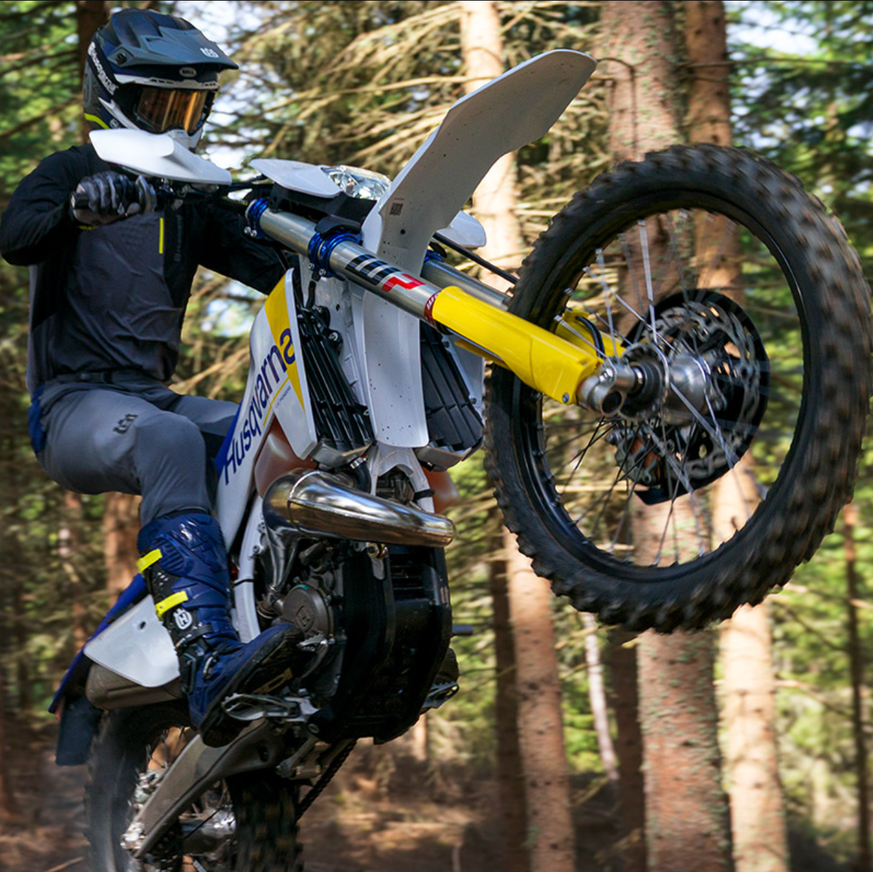 A rider on a white and yellow off-road motorcycle performs a wheelie in a dense pine forest.