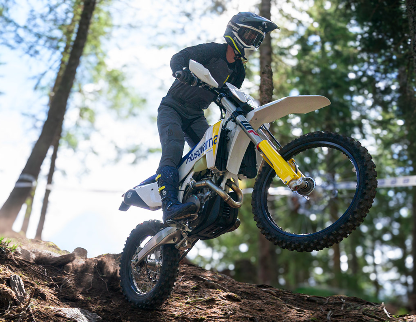 A person riding a white and yellow Husqvarna dirt bike over a dirt track in a forest setting.