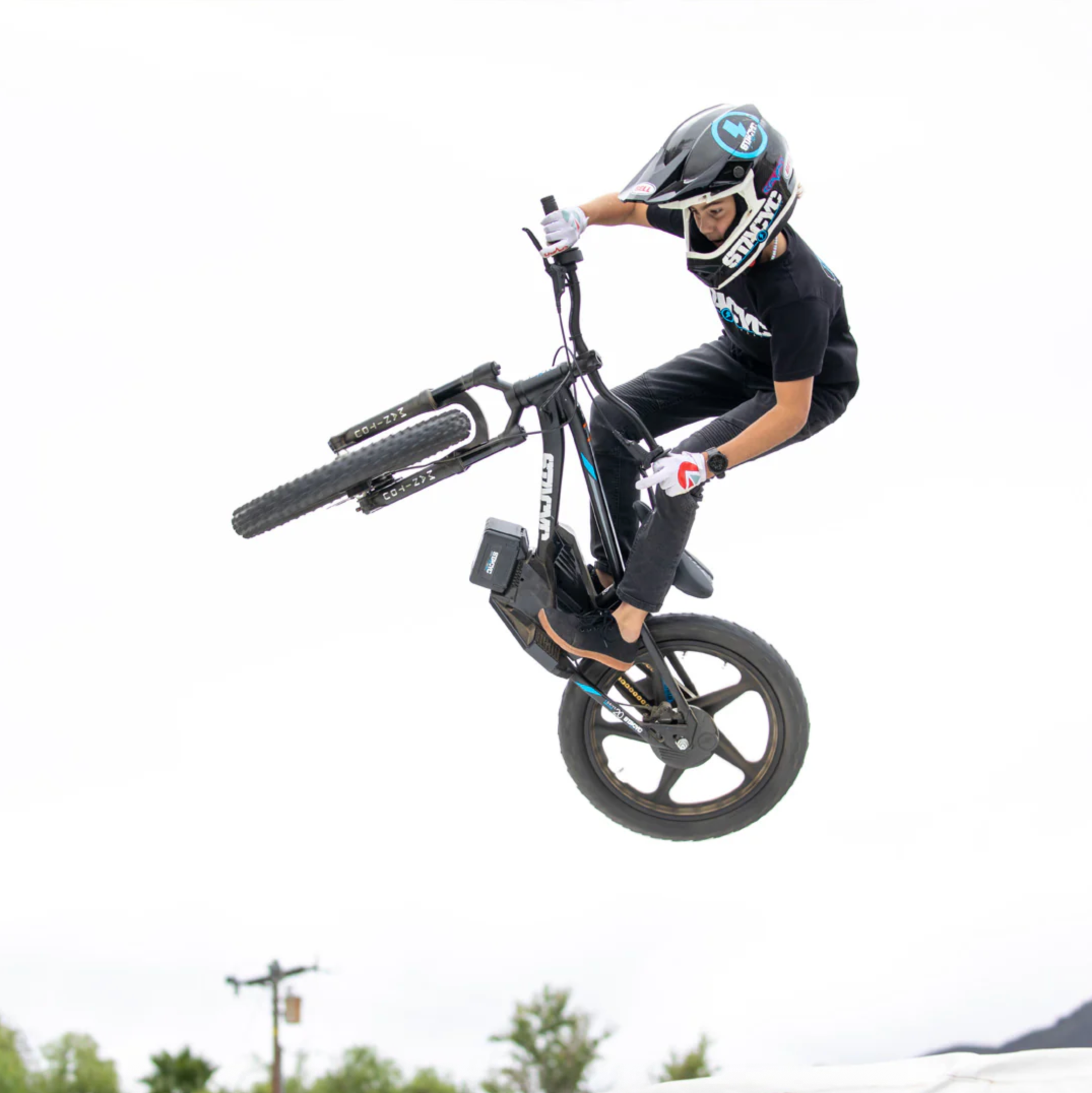 A person in a black outfit and helmet performs a wheelie on a black BMX-style bike against a bright white sky, with a small outdoor scene visible at the very bottom edge.