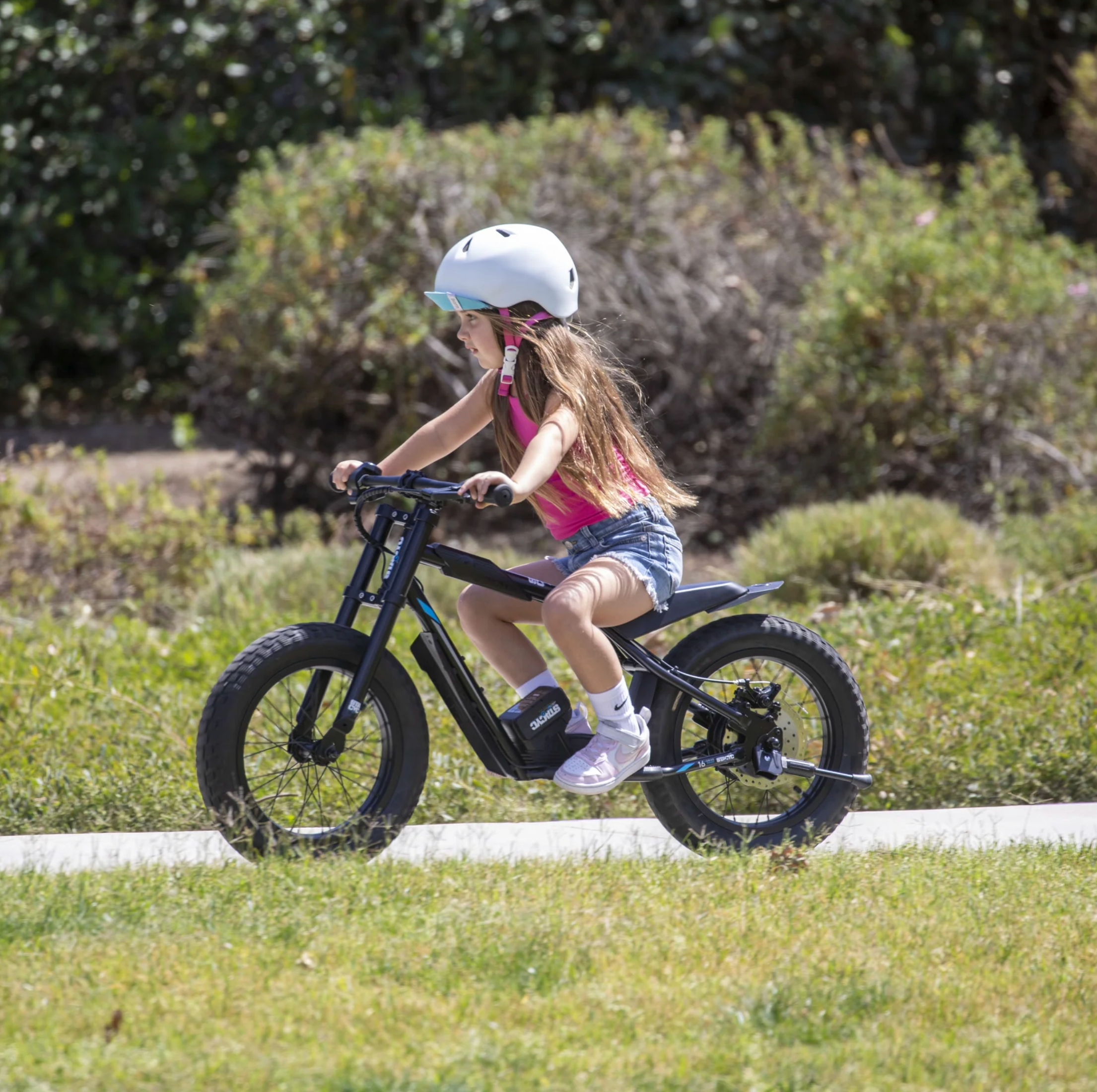 A young girl wearing a white helmet rides a black fat-tire electric balance bike on a paved path in a grassy park with bushes in the background.