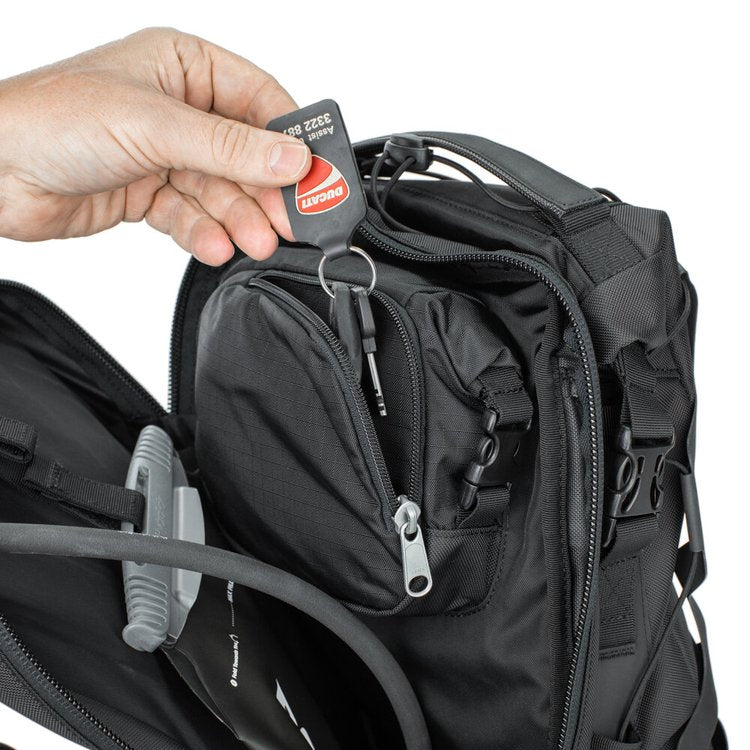 Person holding a key fob above a black backpack with multiple zippers and compartments.